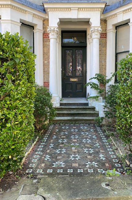 A neat front garden viewed through overhanging tree branches, with a well-maintained lawn bordered by lush, trimmed shrubs and flower beds. The garden features a variety of plants, including dense eve